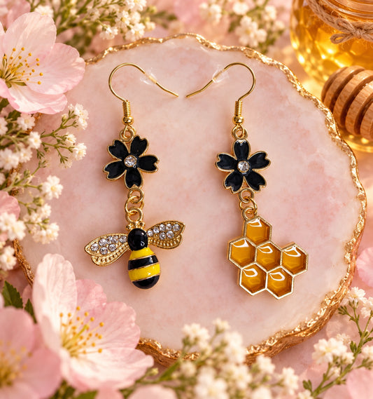 Asymmetric gold-tone earrings with black flowers, a yellow and black bee with sparkling wings, and an amber honeycomb charm.