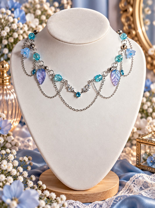 Layered silver-tone necklace with aqua beads, blue flower charms, lavender leaves and a blue centre accent framed by branching details.