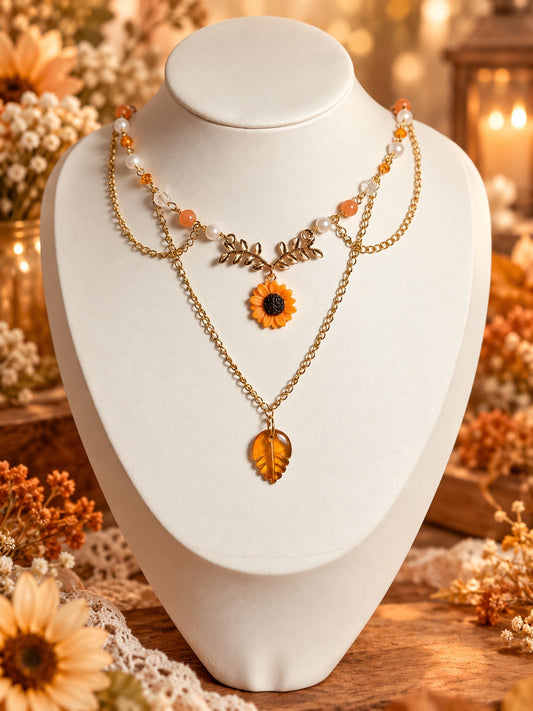 Layered gold-tone necklace with orange and pearly beads, a sunflower beneath leafy branches and an amber-coloured leaf pendant.