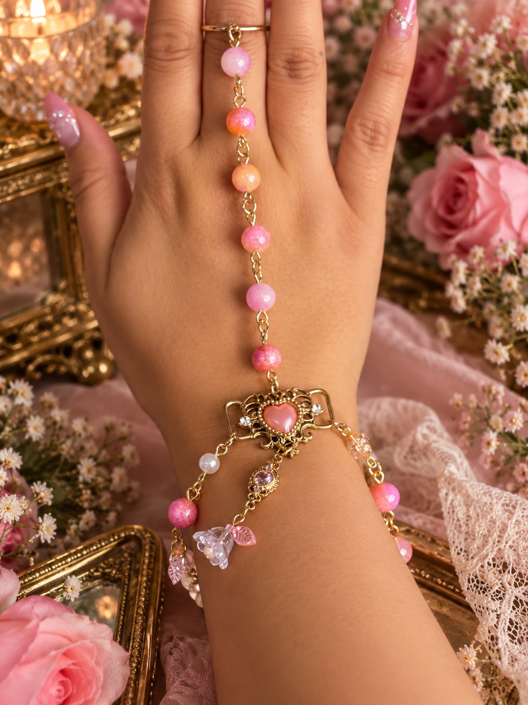 Pink and peach beaded gold-tone handchain with an ornate heart, flower and leaf charms, worn on the hand.