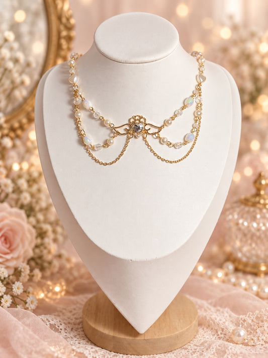 Angelcore gold-tone necklace with an ornate winged heart centrepiece, ivory faux pearls, iridescent and clear beads, and draped chains