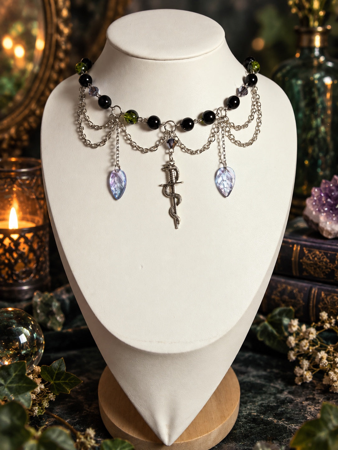 Silver-tone layered necklace with black and olive-green beads, pale purple leaf charms and a serpent wrapped around a dagger pendant.