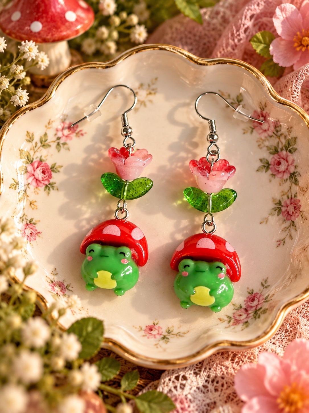 Cottagecore earrings with pink flowers, green leaves and smiling green frogs wearing red spotted mushroom caps